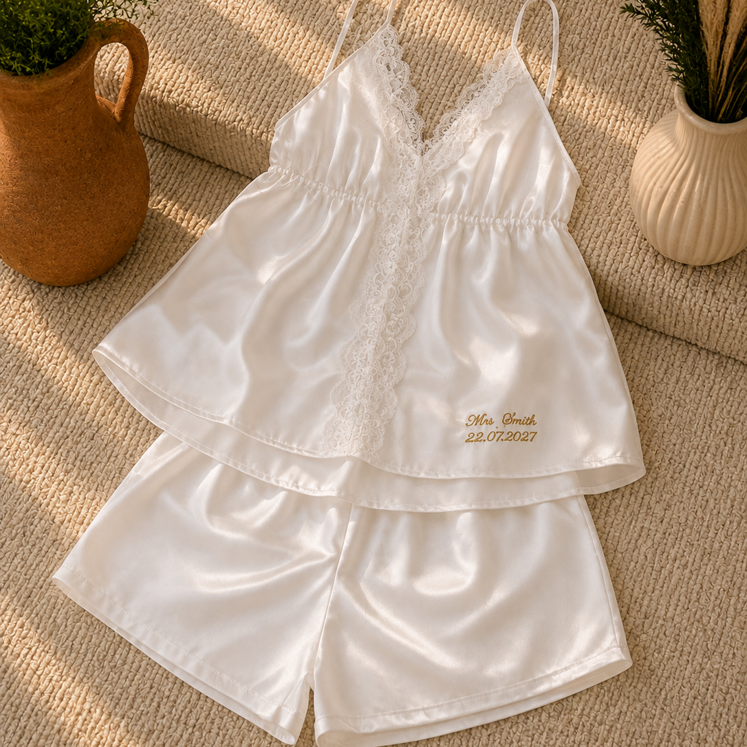 A two-piece white satin pajama set with lace trim, featuring a camisole top and matching shorts, laid flat on a textured beige rug with decorative potted plants.