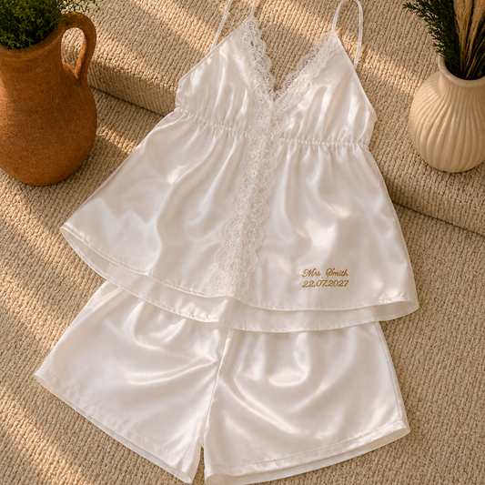 A two-piece white satin pajama set with lace trim, featuring a camisole top and matching shorts, laid flat on a textured beige rug with decorative potted plants.