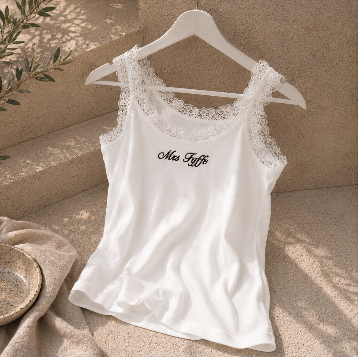 A white camisole tank top with lace shoulder straps and black cursive text reading 'Mes Dits' on the chest, hanging on a white hanger against a beige textured wall with soft natural shadows.
