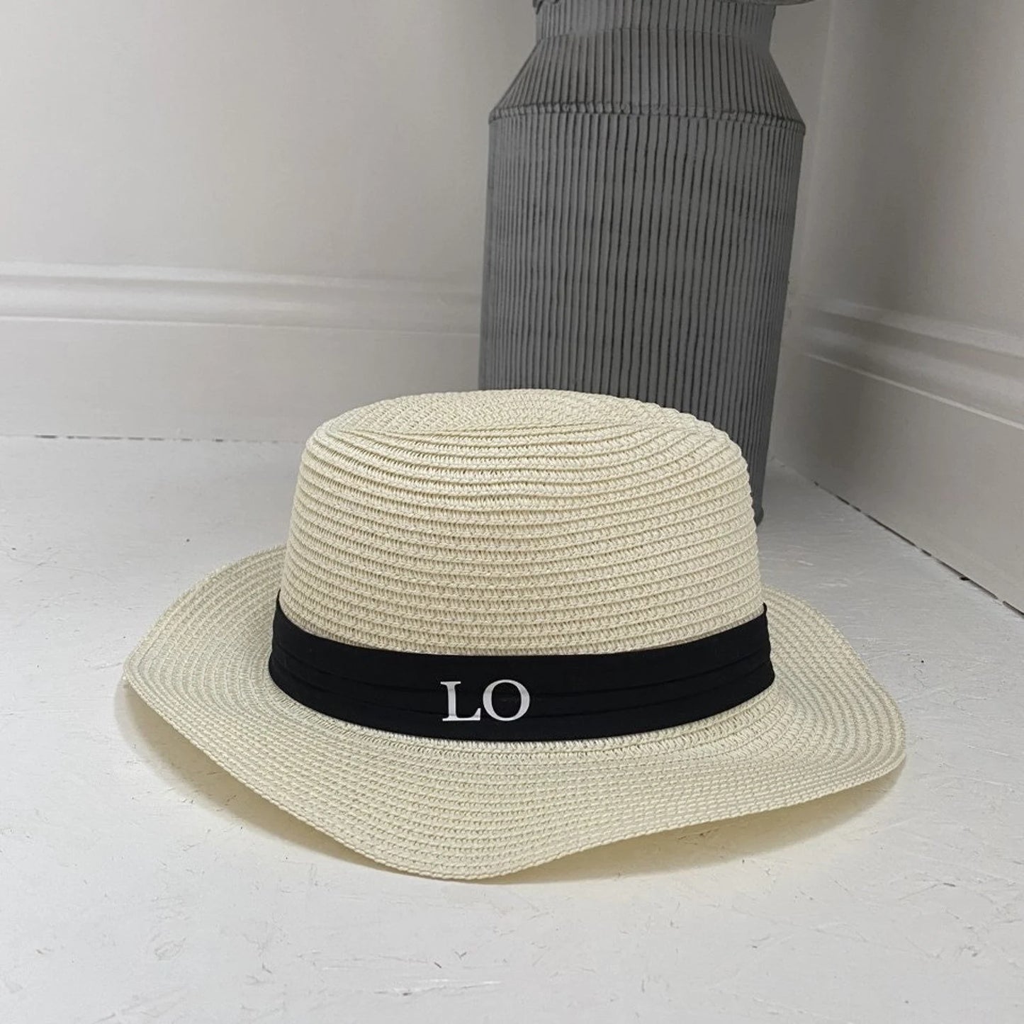 Beige straw hat with a black band featuring 'LO' on a white surface.