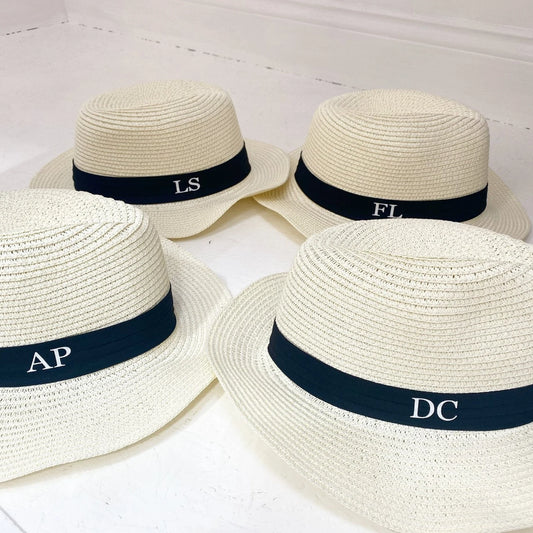 Four straw hats with black bands and monogrammed letters on a white background