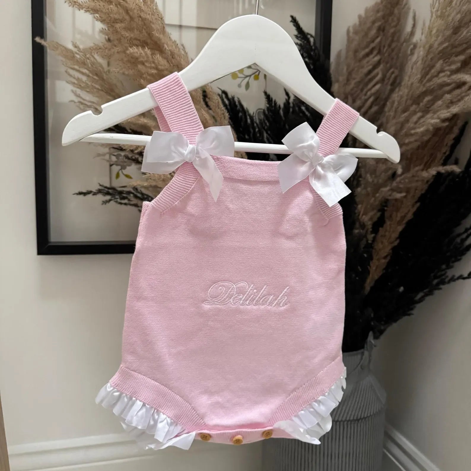 Pink baby romper with ruffles and bows on a hanger against a neutral wall.