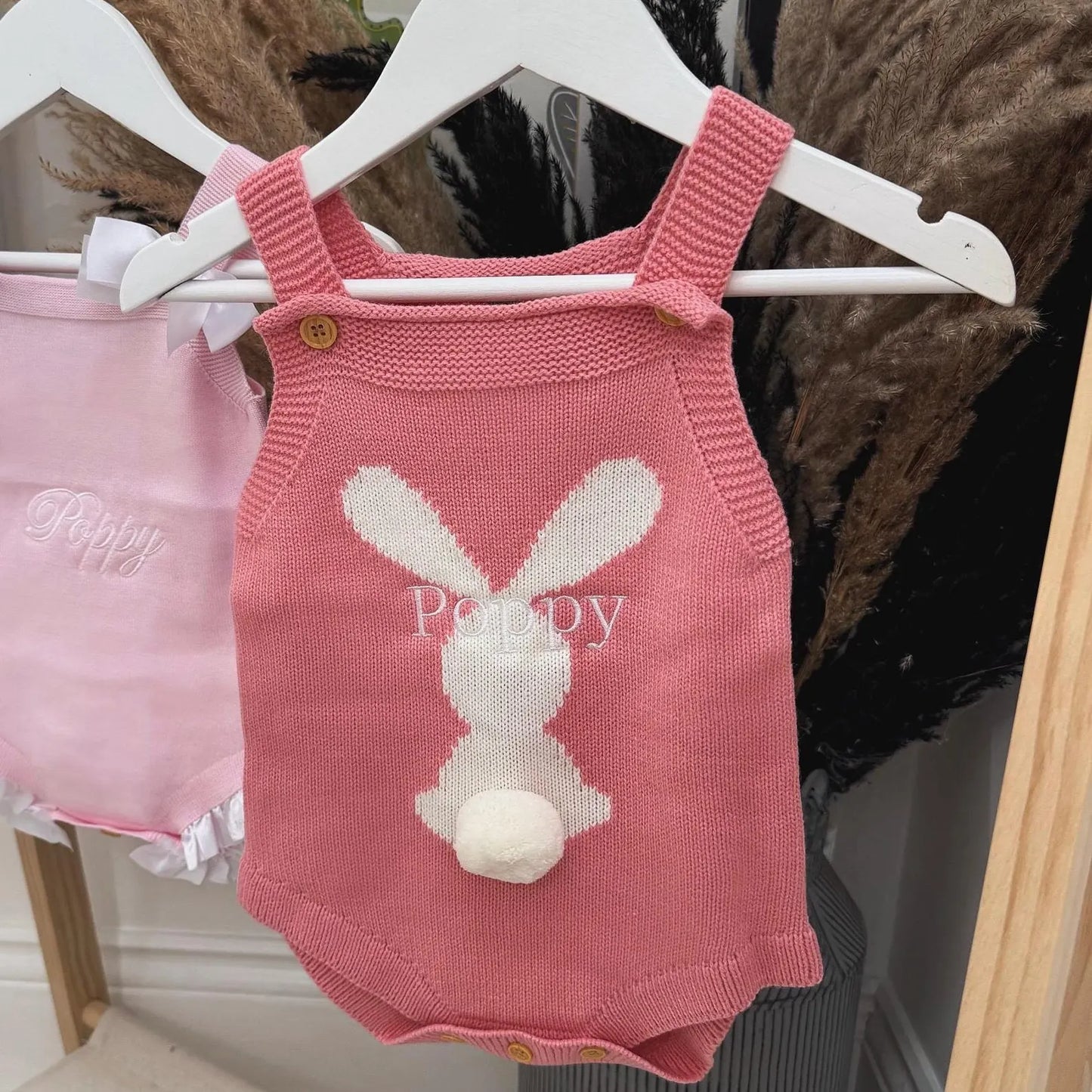 Pink knitted baby romper with bunny design on a hanger against a dark background.