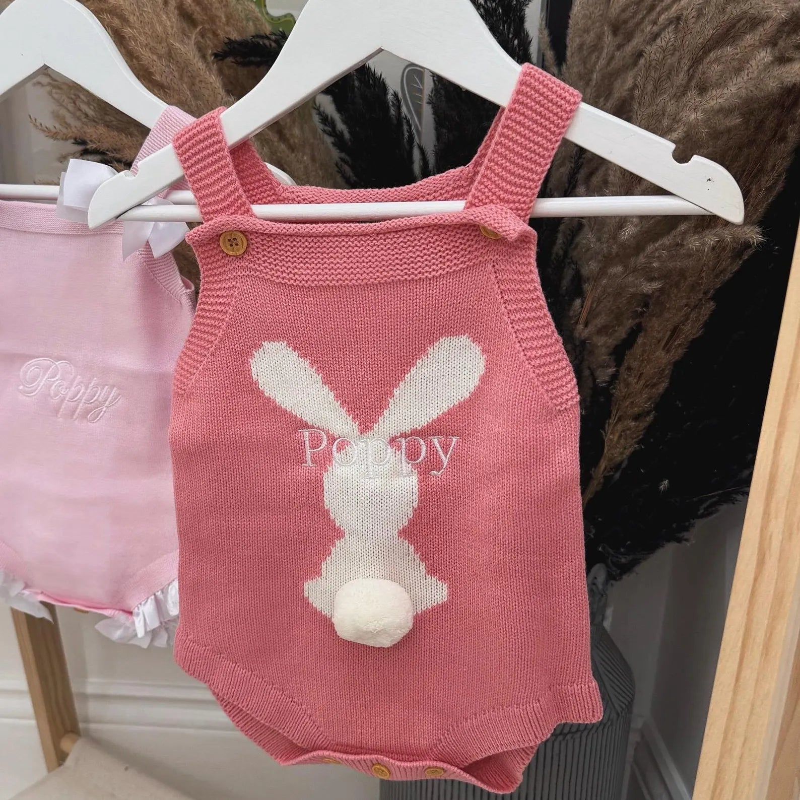 Pink knitted baby romper with bunny design on a hanger against a dark background.
