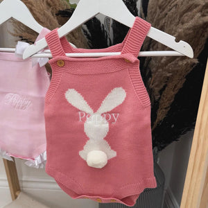 Pink knitted baby romper with bunny design on a hanger against a dark background.