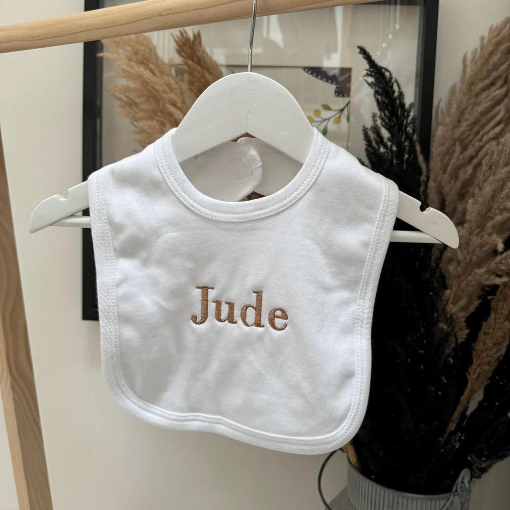 White baby bib with 'Jude' embroidered on it, hanging on a wooden rack.