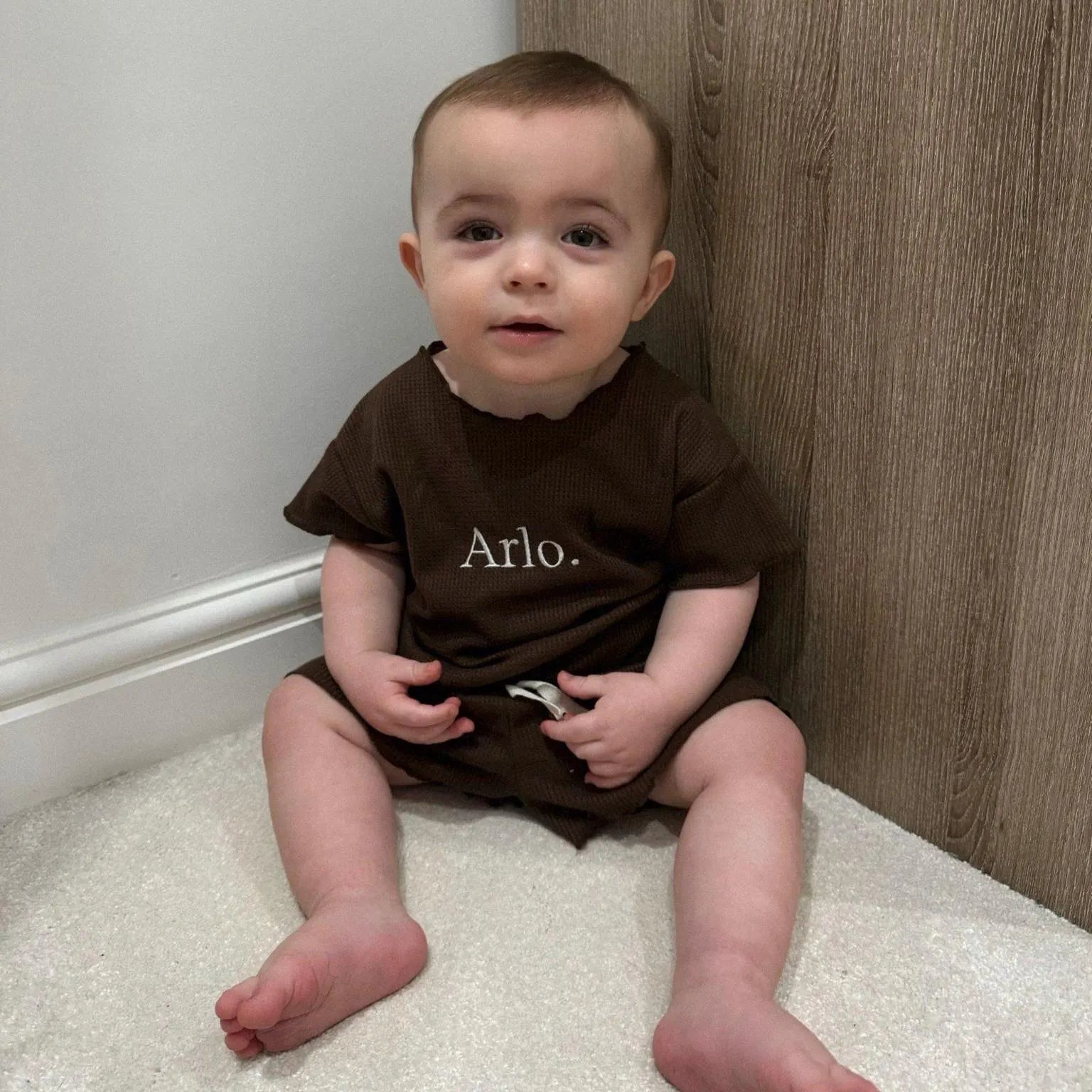 Baby wearing a brown outfit with 'Arlo' printed on it, sitting on a carpeted floor.