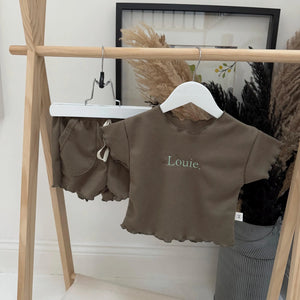 Children's brown outfit with 'Louie' on a hanger against a white wall.