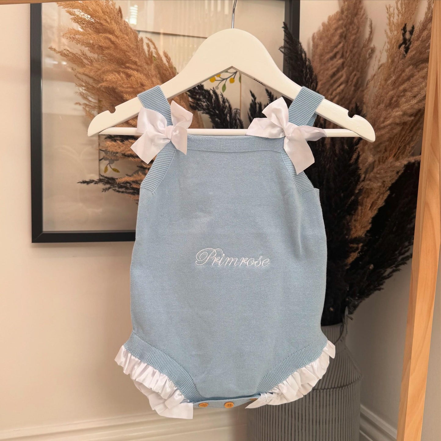 Blue baby romper with white ruffles and bows on a hanger against a neutral wall with personalisation