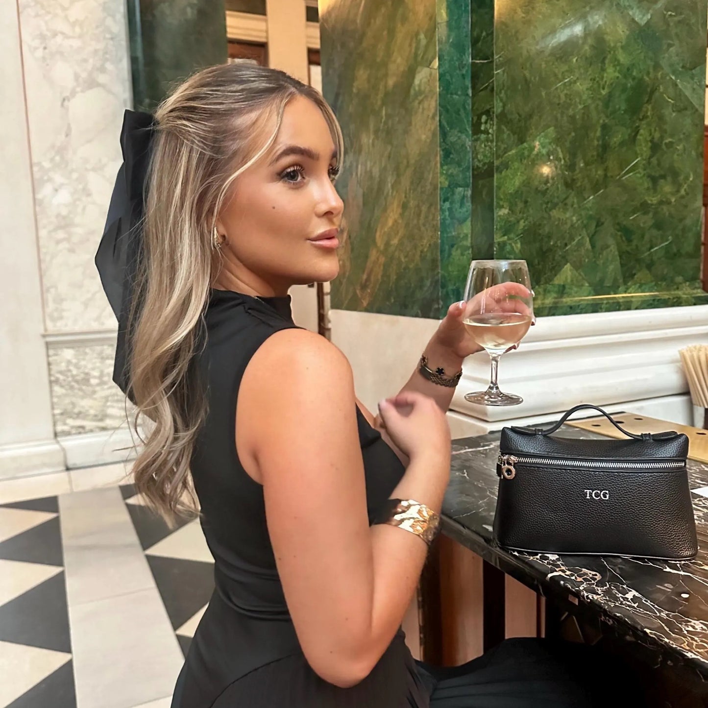 Woman in a black dress holding a glass of wine in an elegant interior setting with black personalised bag