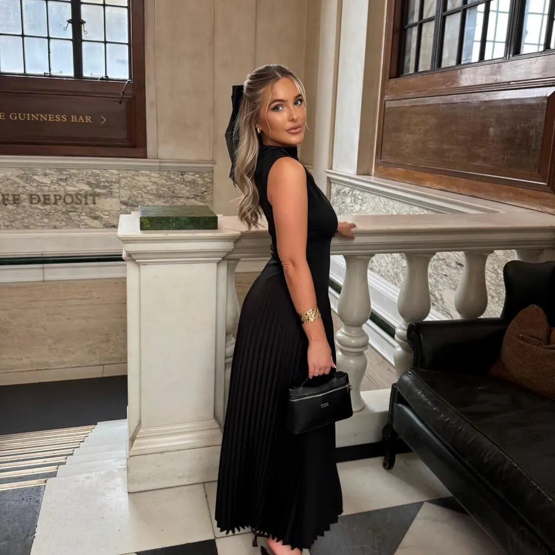 Woman in a black dress standing in an elegant interior setting with marble walls and wooden doors. Holding a black personlised bag