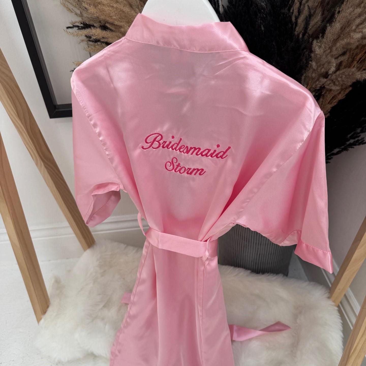 Pink satin robe with 'Bridesmaid Storm' embroidery on a white surface.