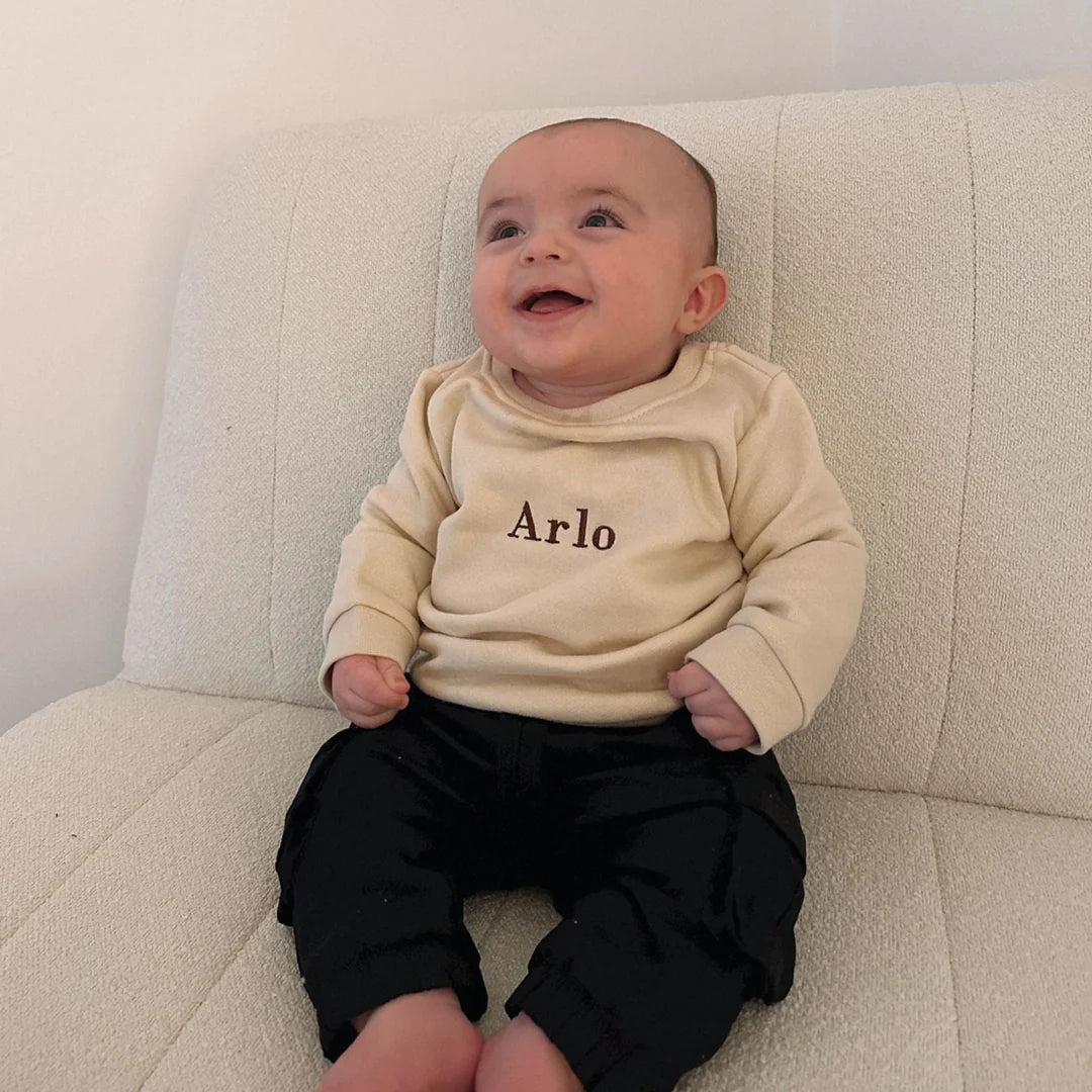 Baby wearing a beige sweatshirt with 'Arlo' on it, sitting on a light-colored couch.