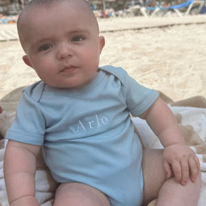 Baby in a light blue onesie with 'Arlo' printed on it, sitting on a sandy beach.