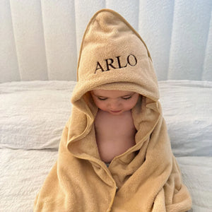 Child wrapped in a beige hooded towel with 'ARLO' embroidered on it, sitting on a white surface.