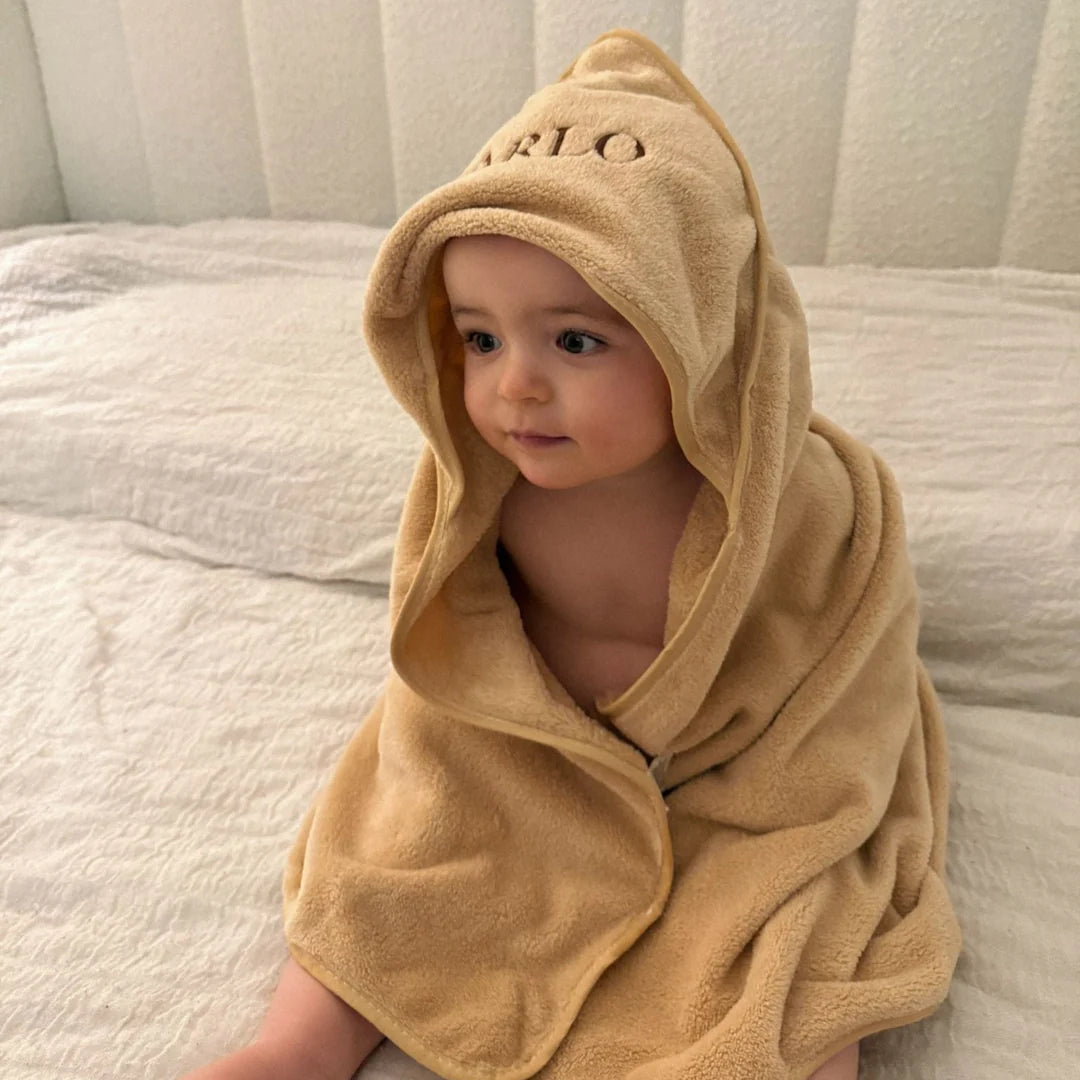 Child wrapped in a beige hooded towel with 'ARLO' embroidery on a bed.