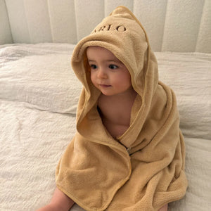 Child wrapped in a beige hooded towel with 'ARLO' embroidery on a bed.