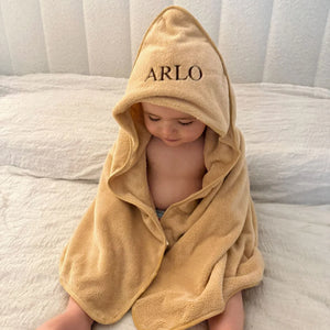 Child wearing a beige hooded towel with 'ARLO' embroidered on it, sitting on a bed.