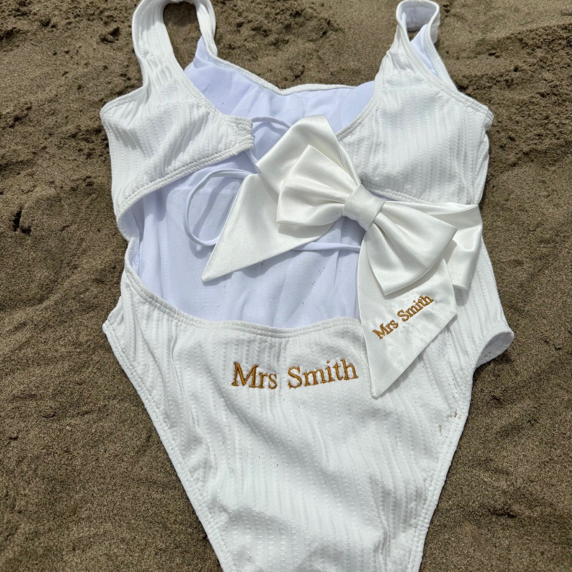 White swimsuit with a large bow and 'Mrs Smith' embroidery on sand