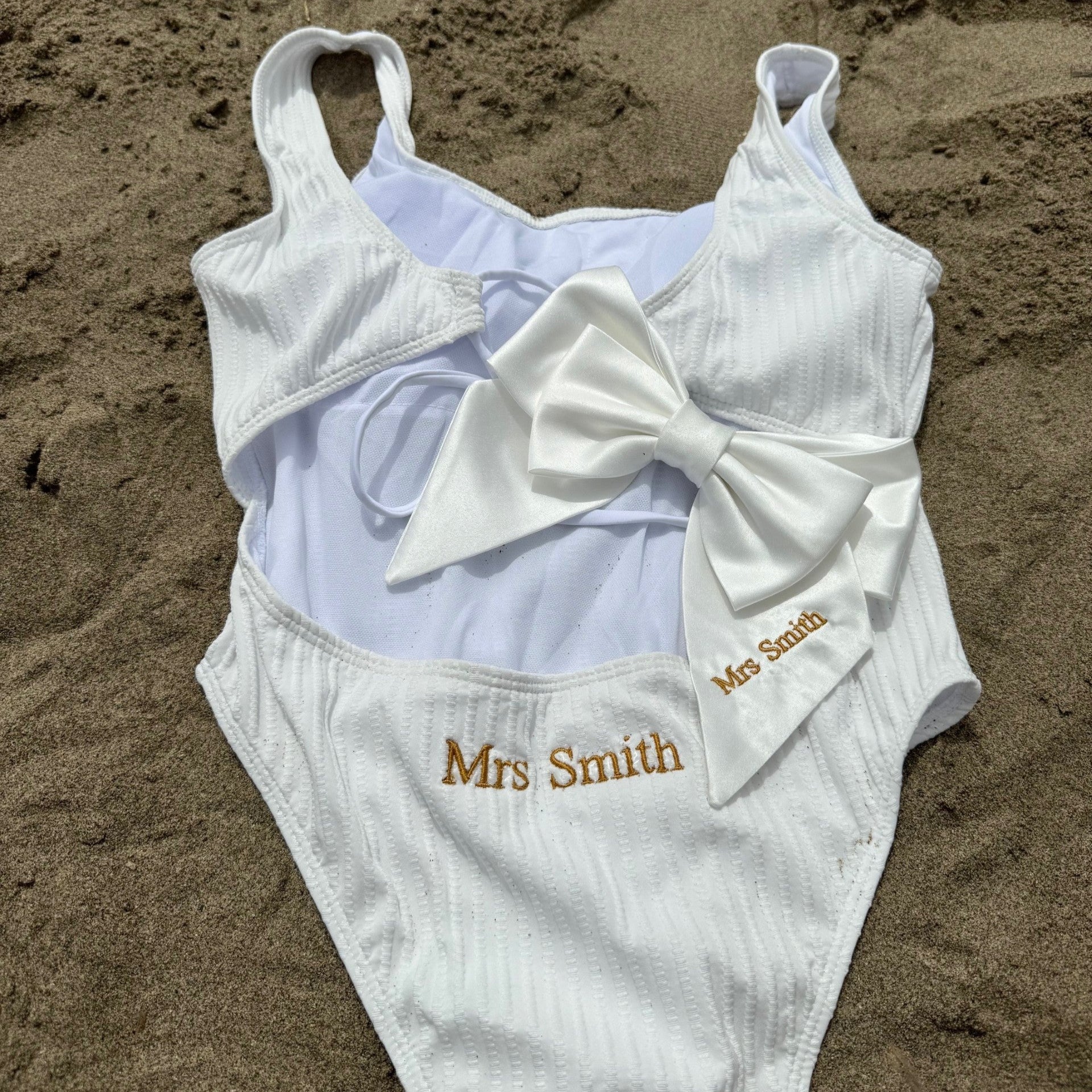 White swimsuit with a large bow and 'Mrs Smith' embroidery on sand