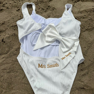 White swimsuit with a large bow and 'Mrs Smith' embroidery on sand