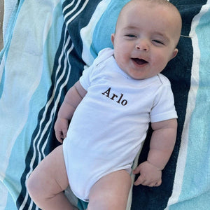 Baby wearing a white onesie with 'Arlo' embroidered on it, sitting on a striped blanket.
