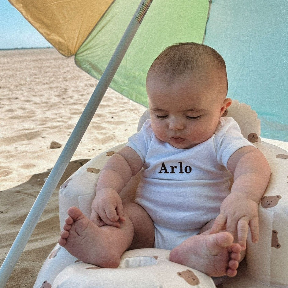 Baby in a white onesie with 'Arlo' on it, sitting in a beach chair under a colorful umbrella.