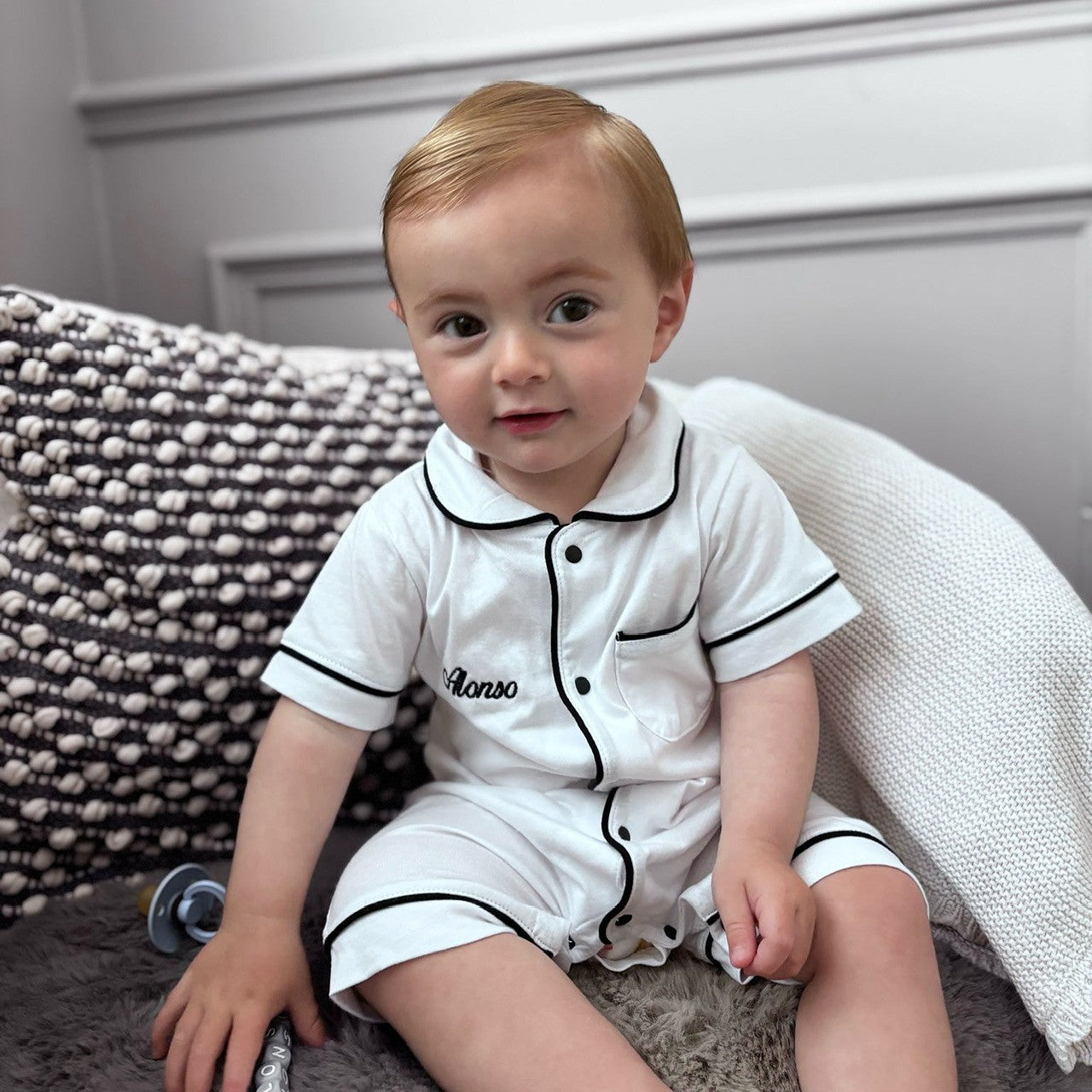 Child wearing a white outfit with black trim sitting on a textured surface.