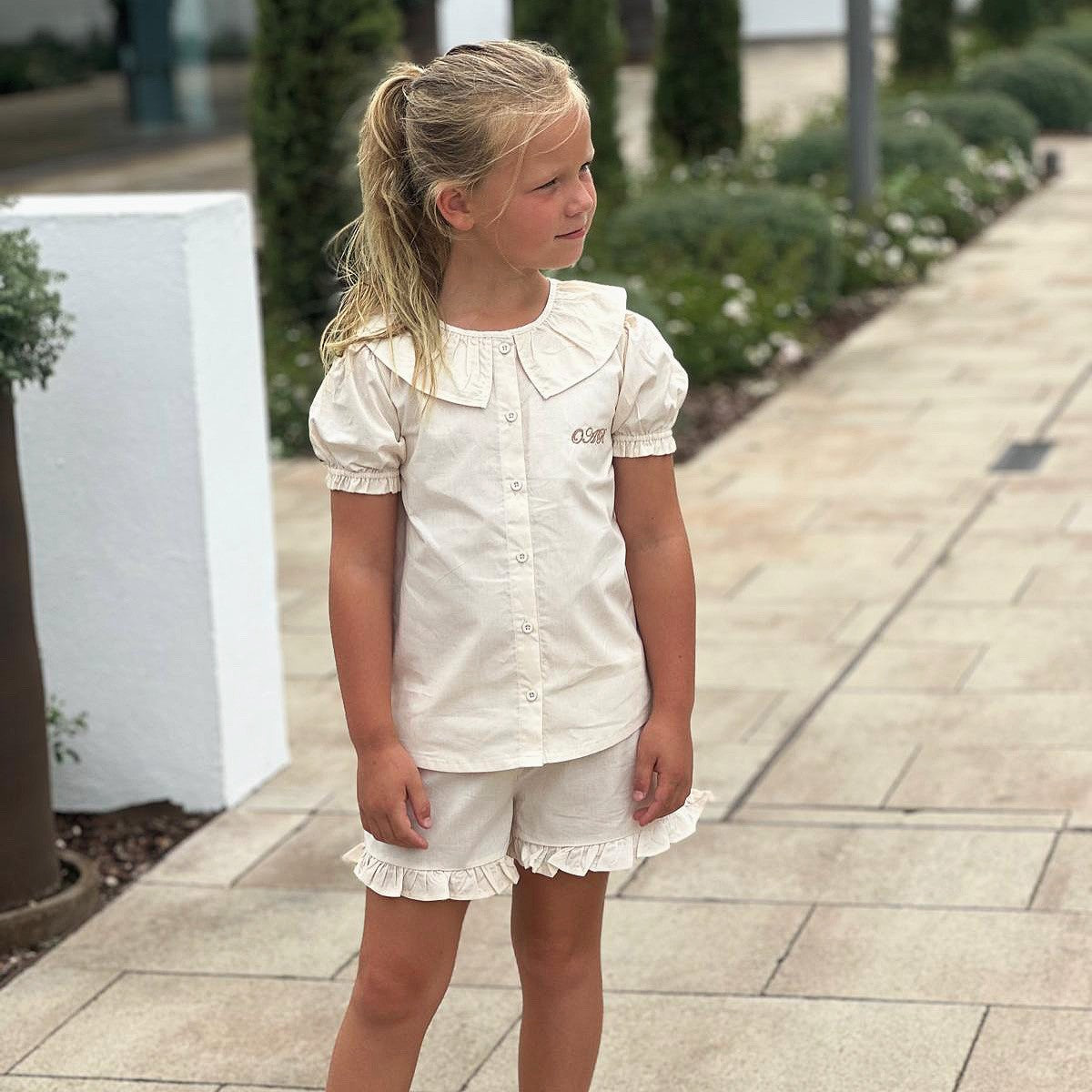 Young girl in a beige outfit with personalisation standing on a paved path with greenery in the background