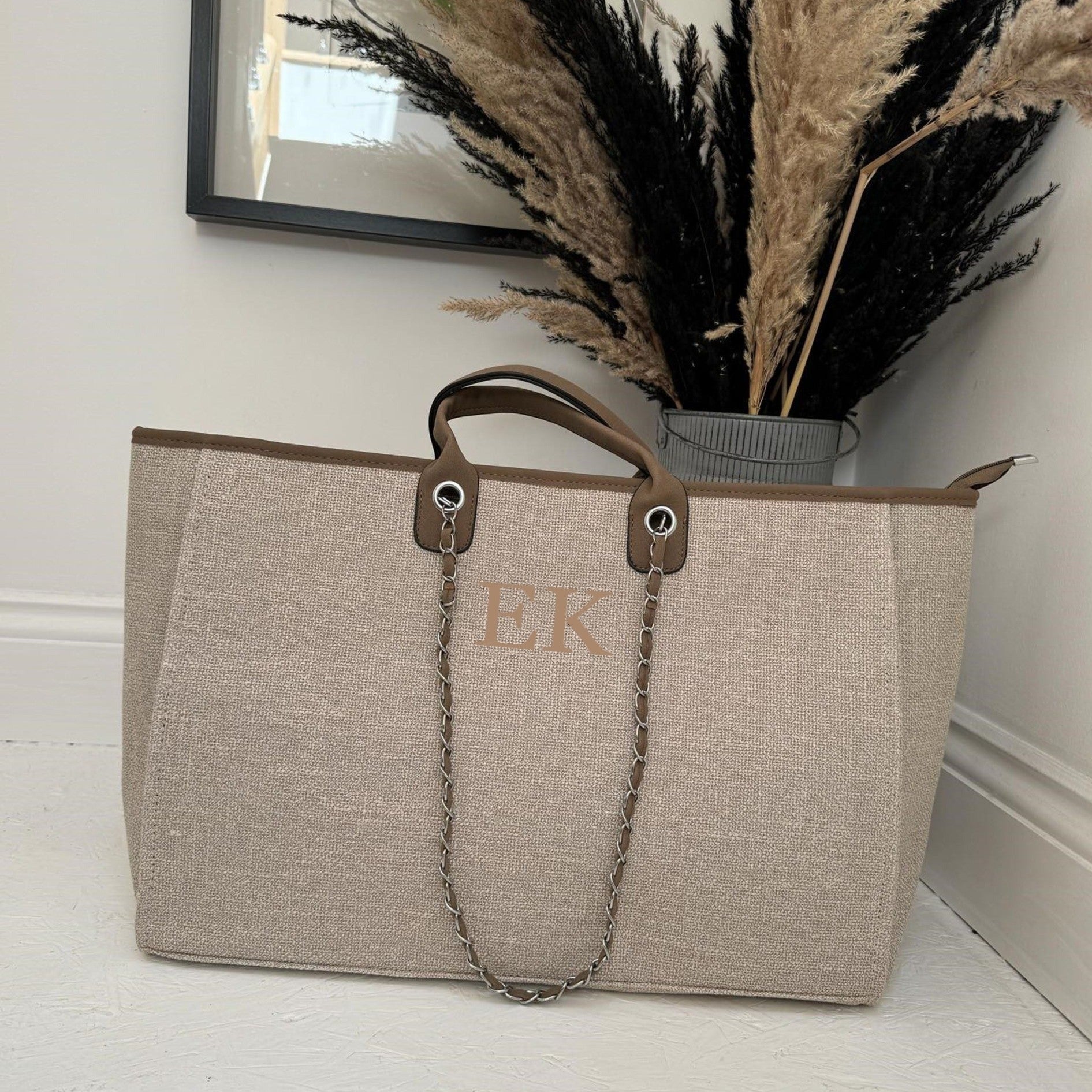 A beige personalised tote bag with a textured surface, a handle, and a chain strap, placed on a floor against a wall with a decorative object and a potted plant in the background.