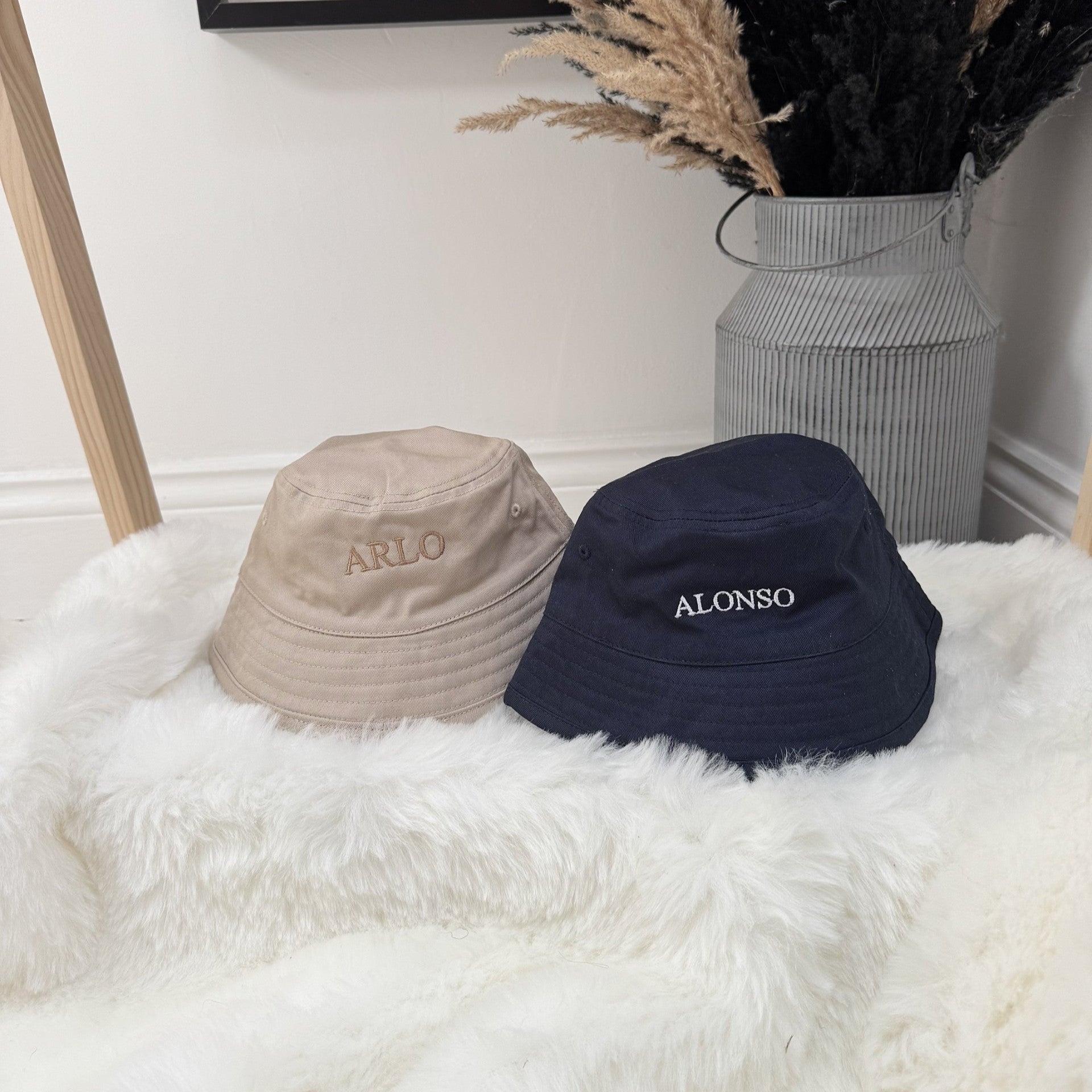 Two bucket hats, one beige and one navy, with 'ARLO' and 'ALONSO' branding, on a white fluffy surface.
