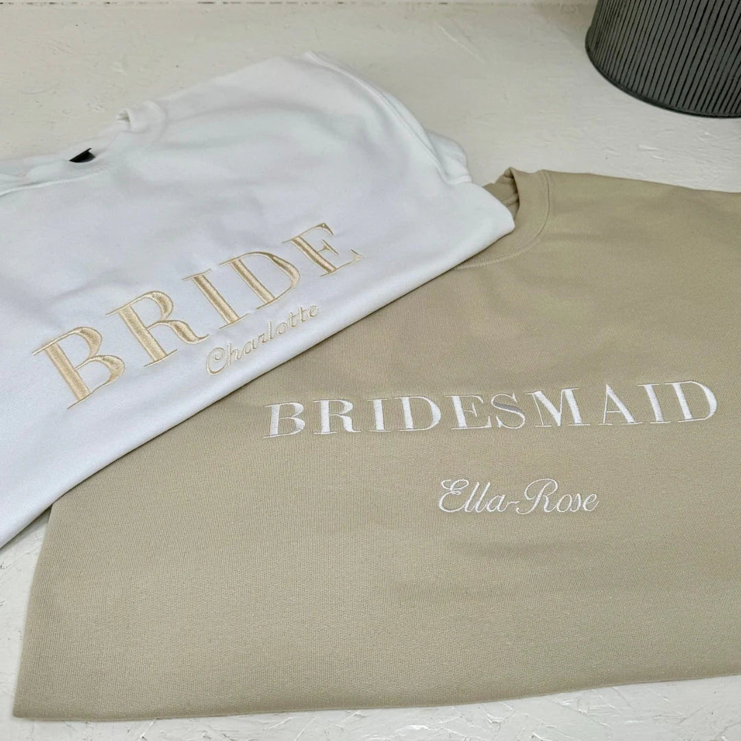 Two sweatshirts, one white with 'BRIDE' and another beige with 'Bridesmaid', on a light surface.