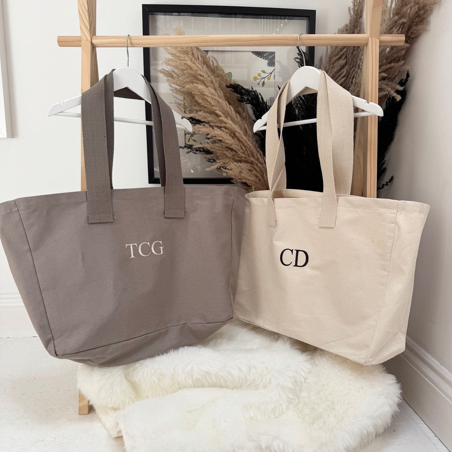 Two tote bags, one gray and one beige, with monogram initials on a wooden stand against a white wall. Both personalised