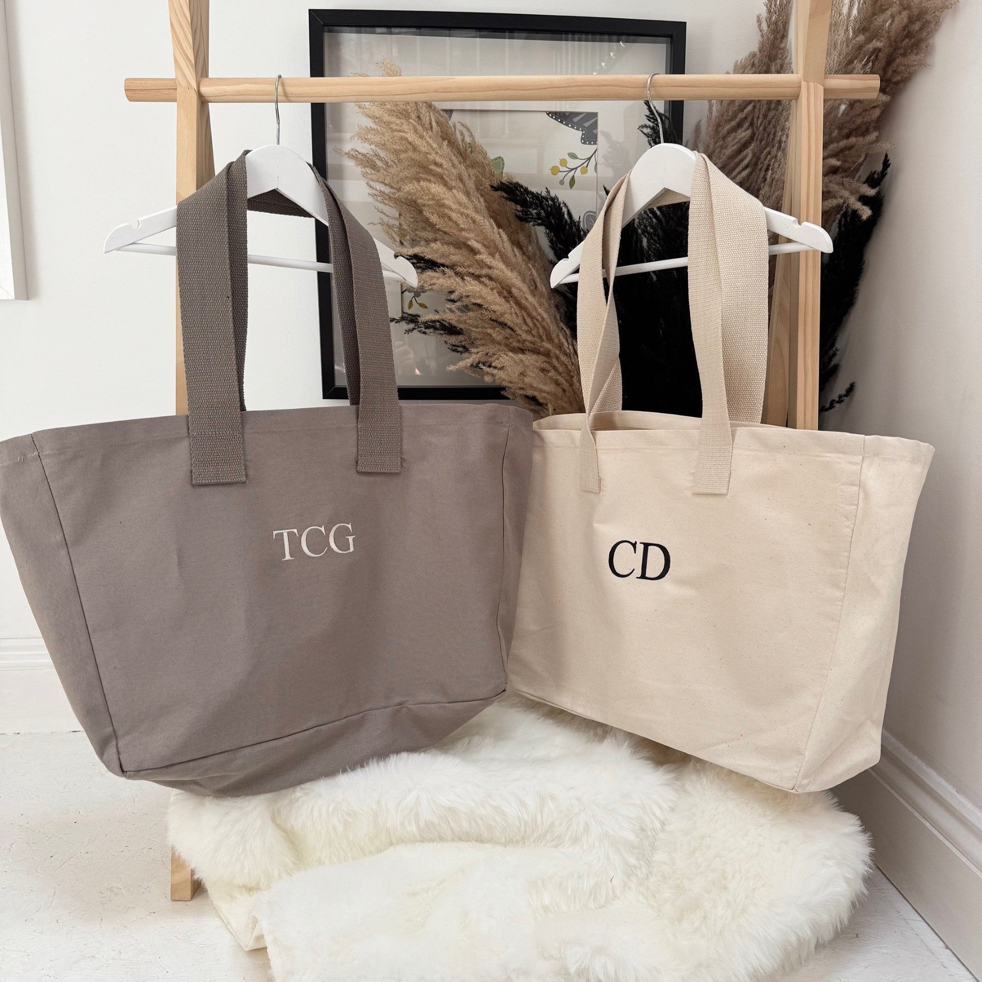 Two tote bags, one gray and one beige, with monogram initials on a wooden stand against a white wall. Both personalised
