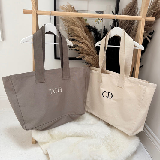 Two tote bags, one gray and one beige, with initials 'TCG' and 'CD', hanging on a wooden rack.