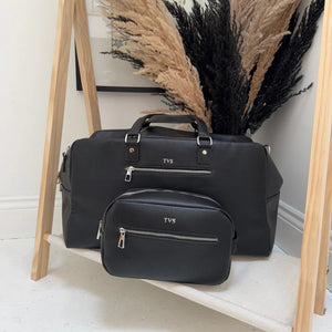 Black duffel bag with 'TVS' branding on a wooden stand against a neutral wall.