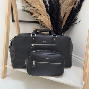 Black duffel bag and smaller pouch with 'TVS' branding on a wooden shelf.