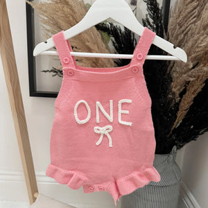 Pink baby romper with 'ONE' and a bow on a hanger against a neutral background