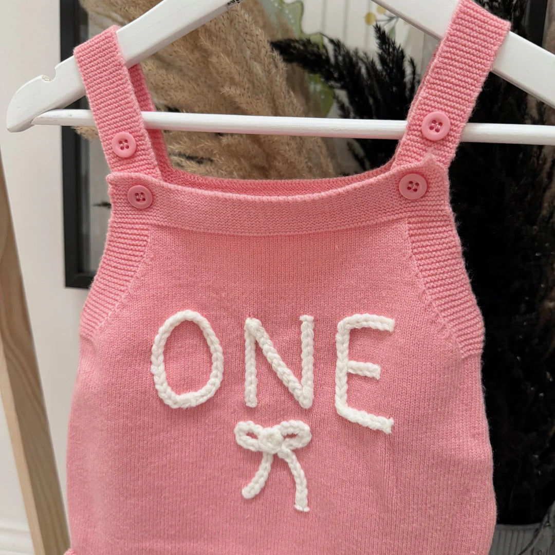 Pink knitted dress with 'ONE' and a bow on a hanger
