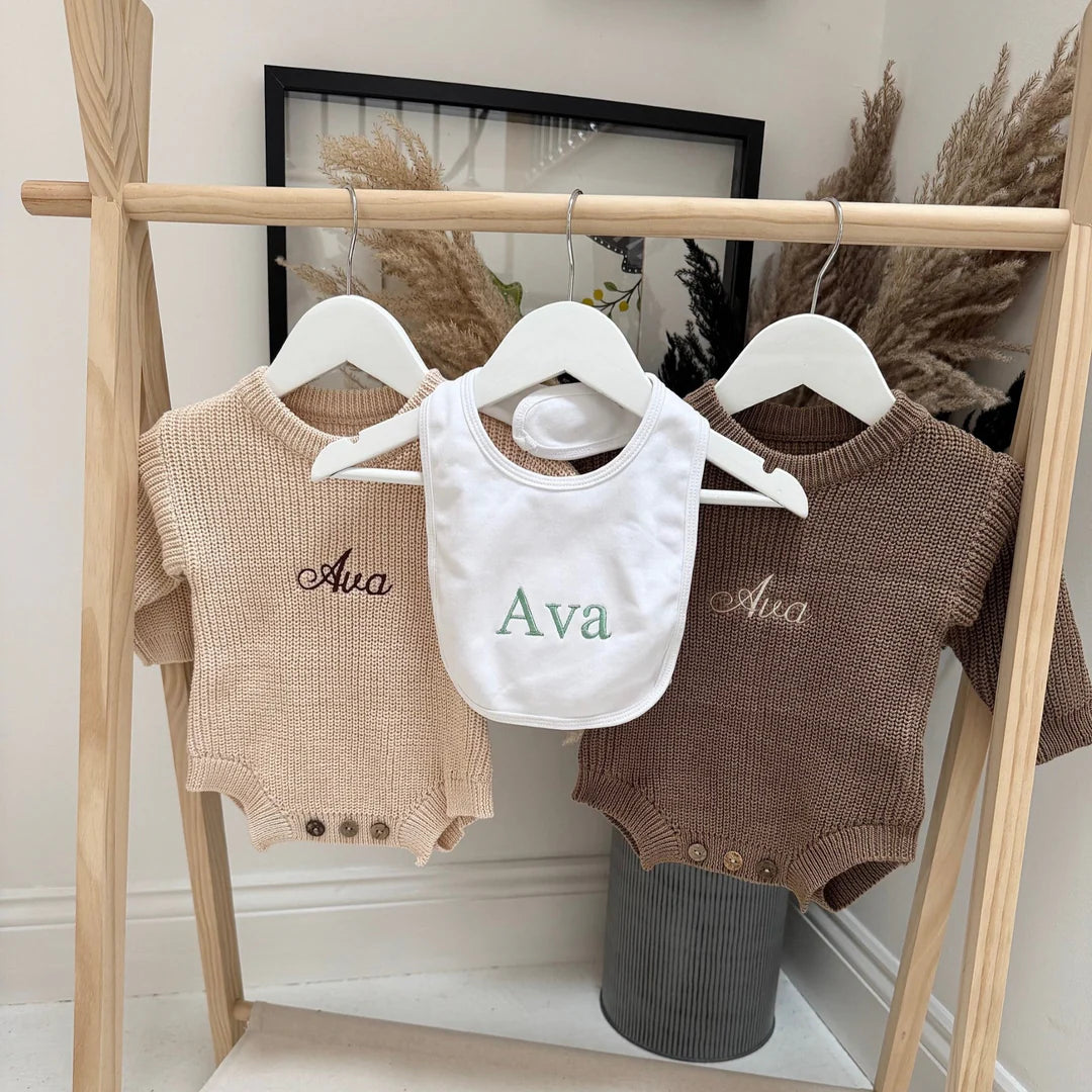 Two brown baby rompers and a white bib with 'Ava' embroidered on them, hanging on a wooden rack.
