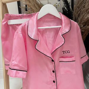 Pink satin pajama set with black trim on a hanger against a neutral background with TCG on
