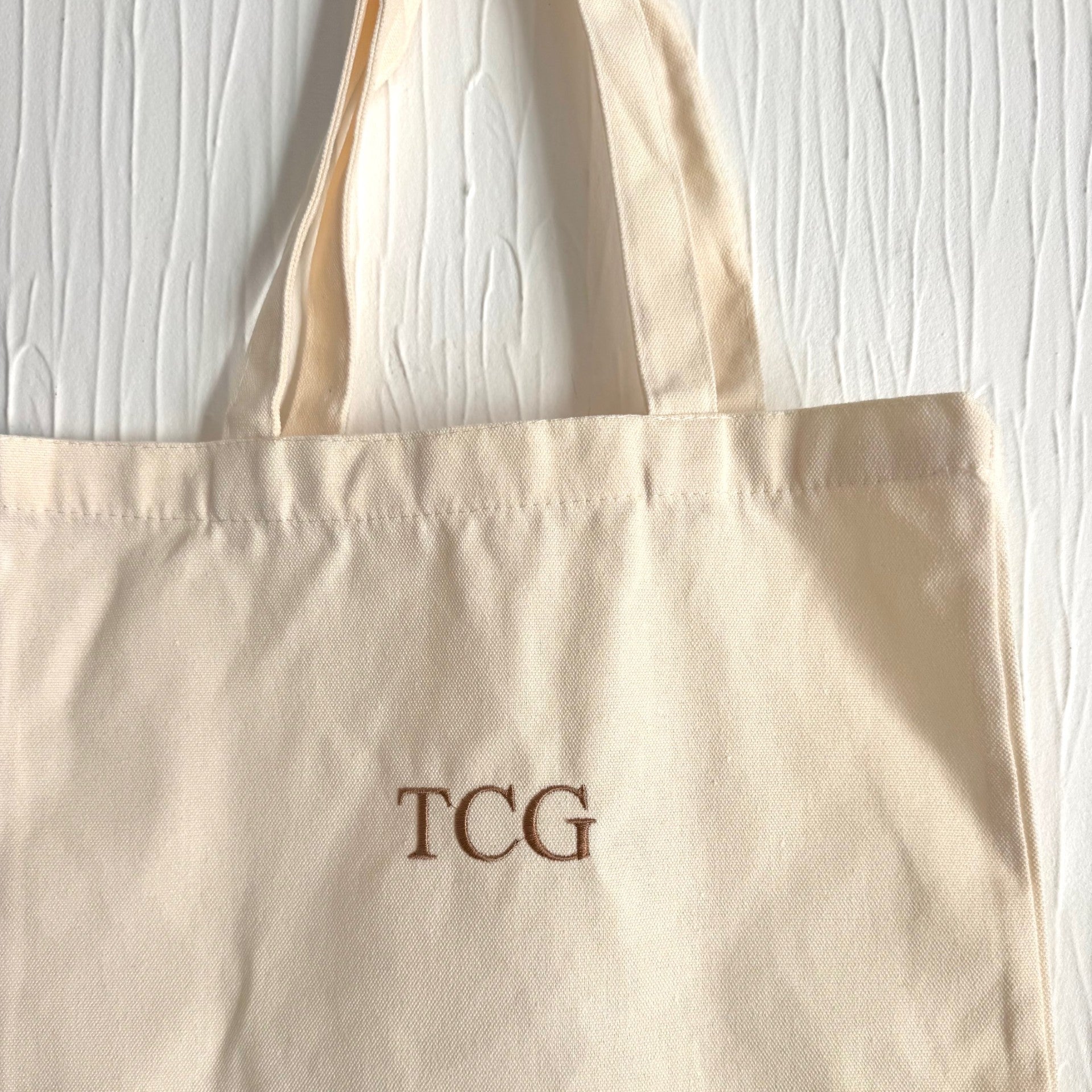Beige tote bag with 'TCG' monogram on a light wooden surface
