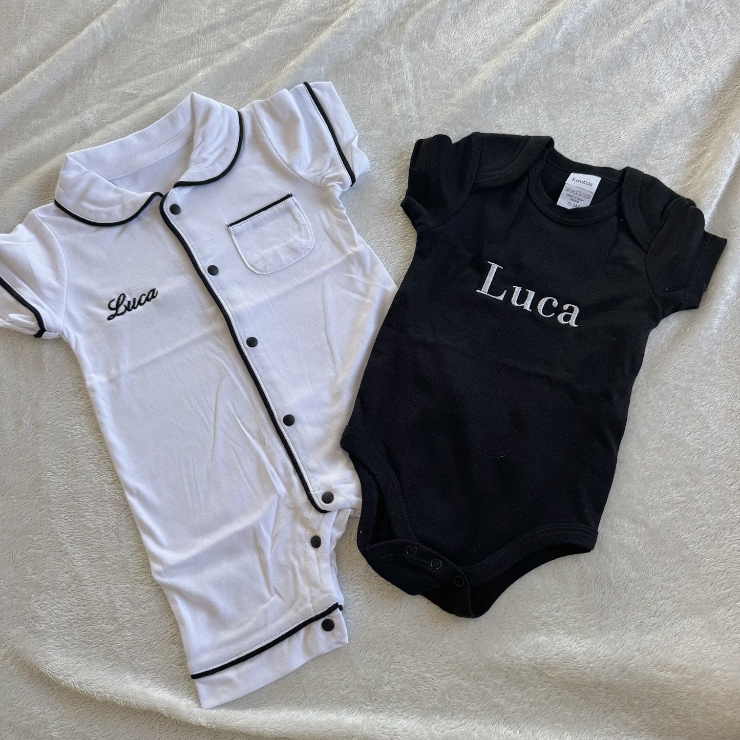 White and black baby rompers with 'Luca' printed on them, laid flat on a textured surface.