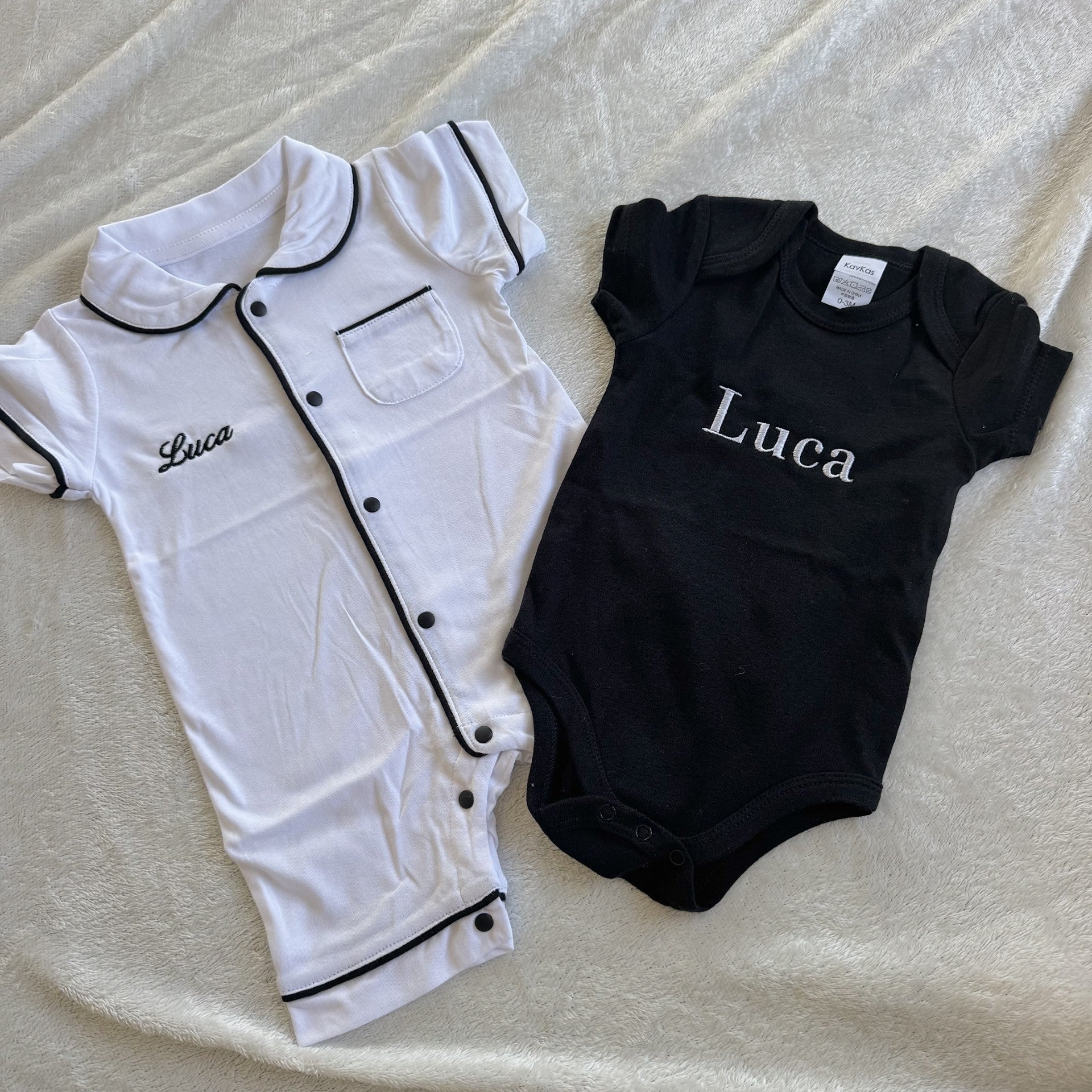 White and black baby rompers with 'Luca' printed on them, laid flat on a textured surface.