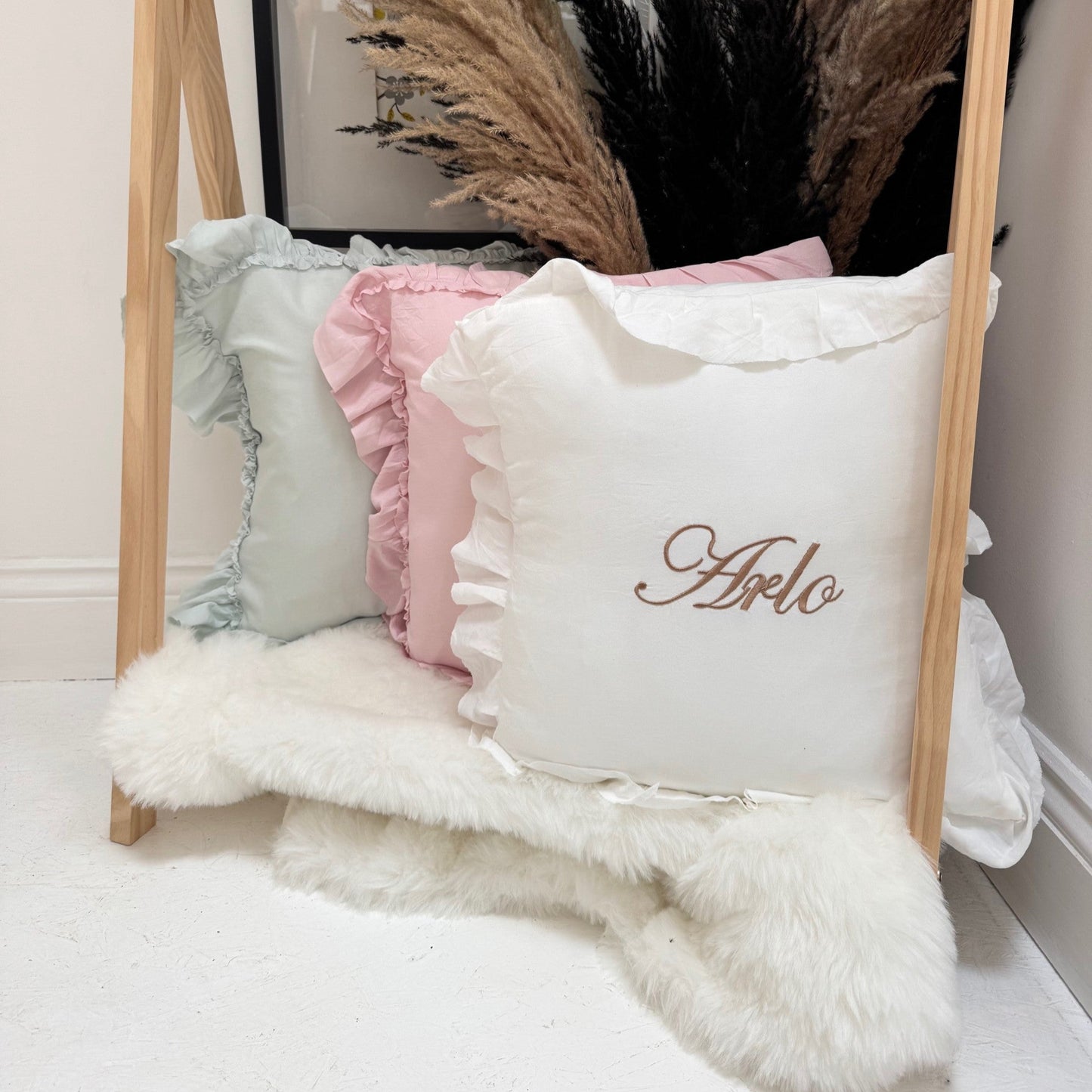 Decorative pillows with ruffles and a personalized name on a wooden stand against a neutral wall.