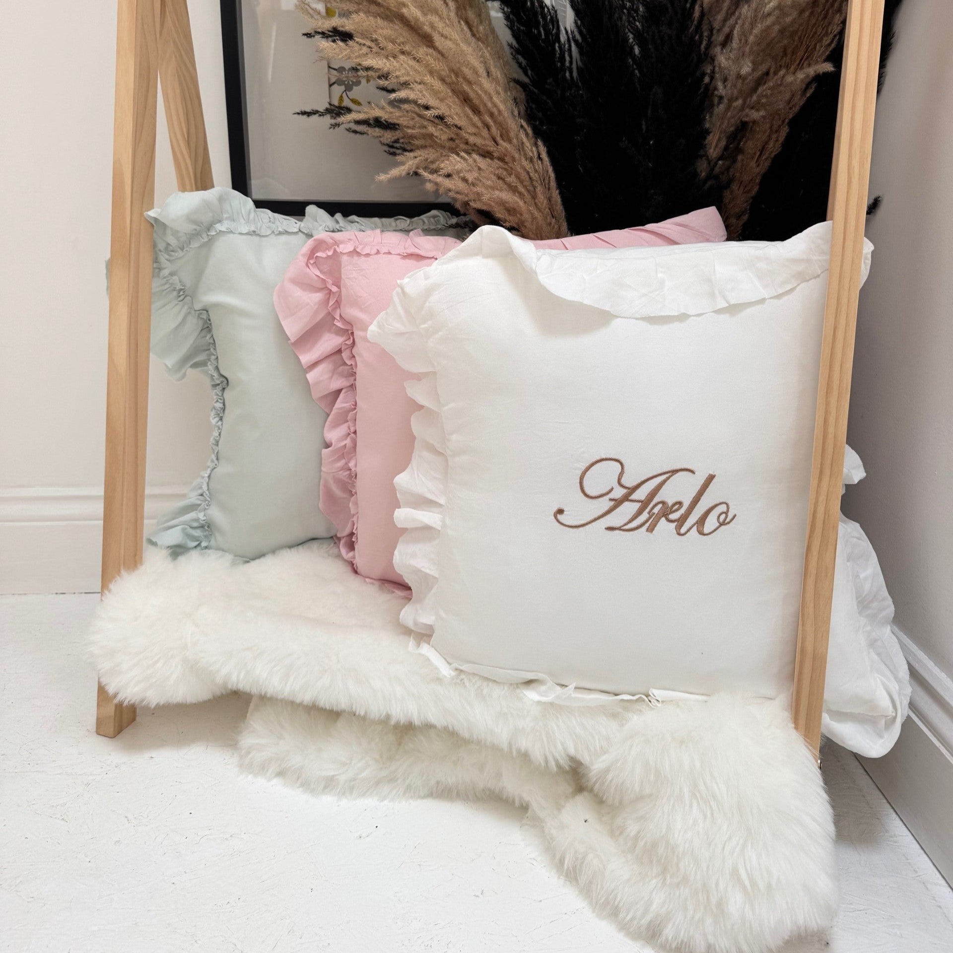 Decorative pillows with ruffles and a personalized name on a wooden stand against a neutral wall.