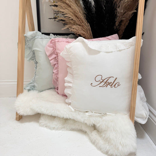 Decorative pillows with ruffles and a personalized name on a wooden stand against a neutral wall.