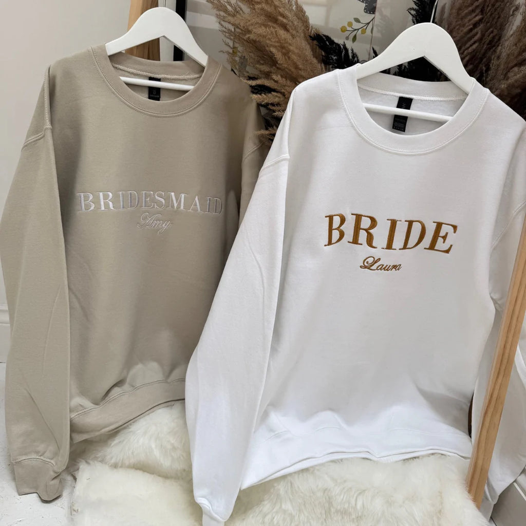 Two sweatshirts, one beige with 'Bridesmaid' and one white with 'Bride', hanging on a wooden rod.