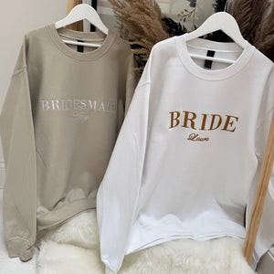 Two sweatshirts, one beige with 'Bridesmaid' and one white with 'Bride', hanging on a wooden rod.