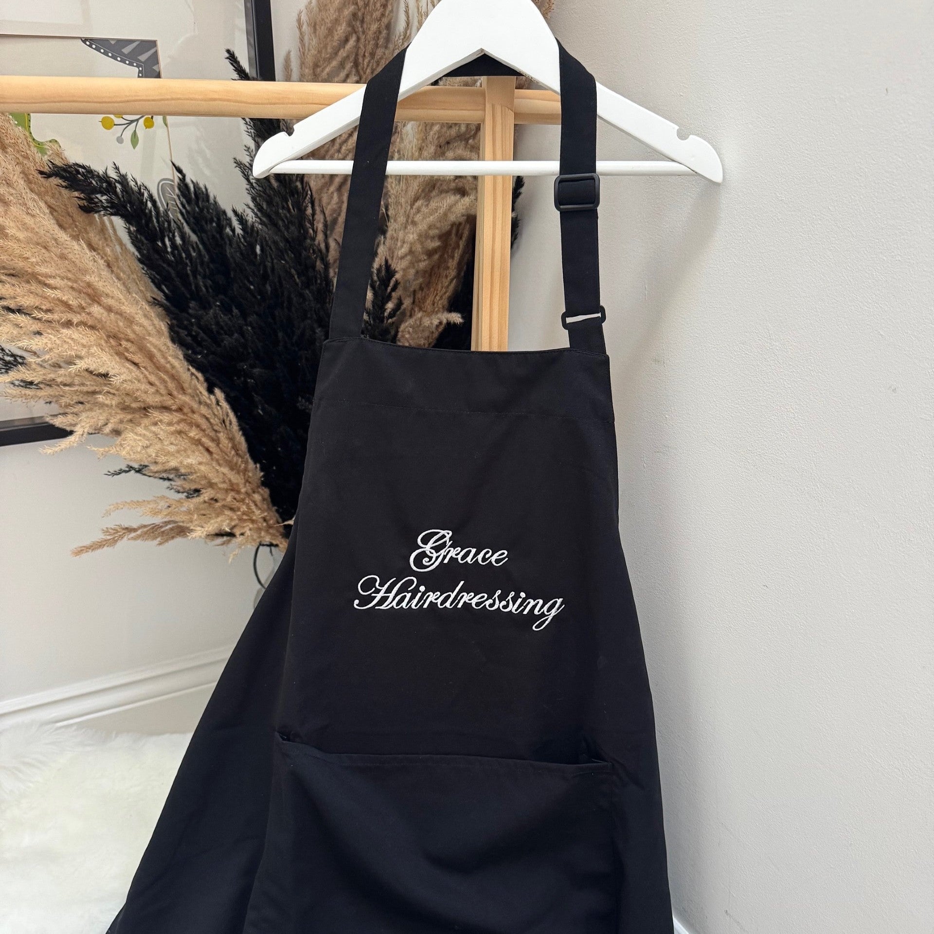 Black apron with 'Grace Hairdressing' text hanging on a white wall.